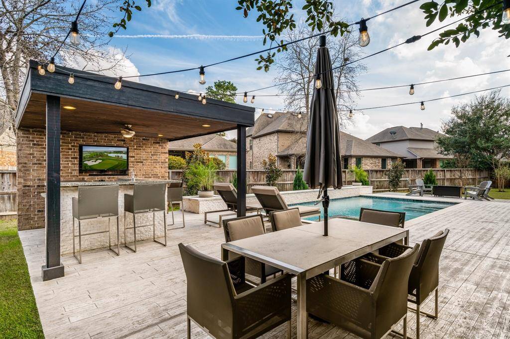 This backyard features a stylish outdoor living area with a covered patio bar, string lights, and a spacious dining setup. A pristine pool is surrounded by stamped concrete that looks like wood, perfect for entertaining and relaxation.