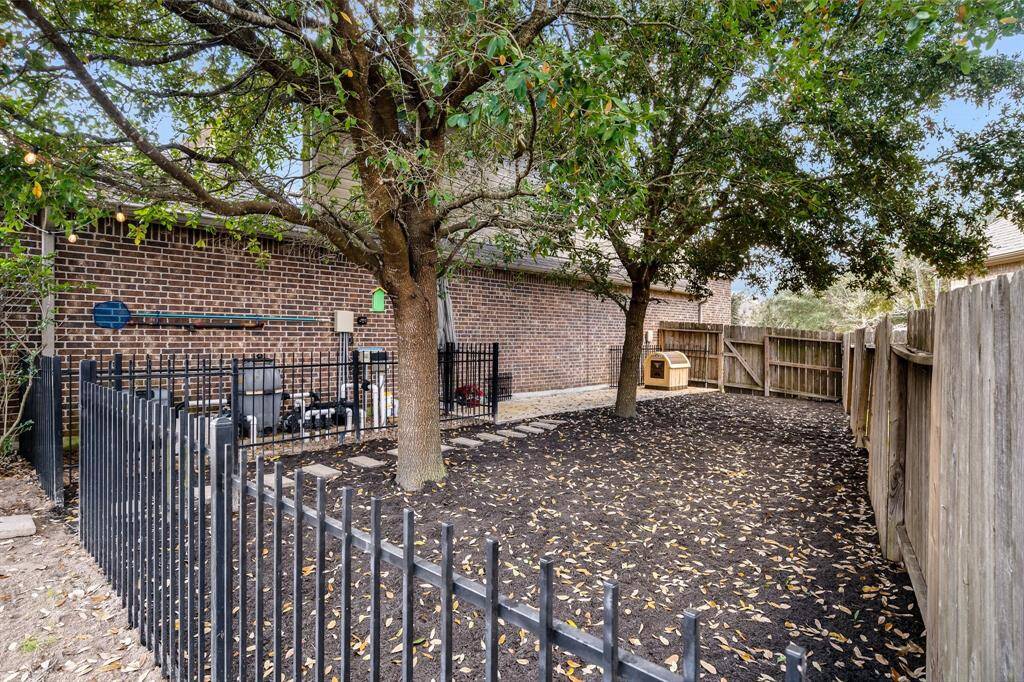 A fenced backyard area with a brick exterior, featuring mature trees providing shade and privacy, a dog house, and a small utility section. Ideal for pets and outdoor activities.
