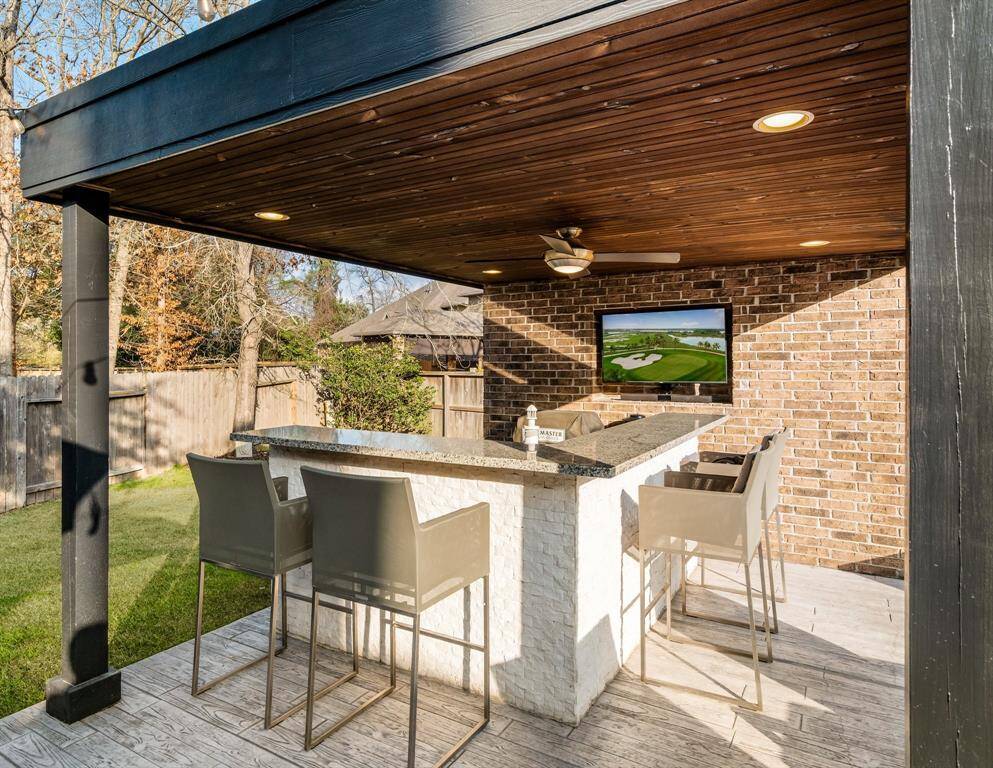 This photo showcases a stylish outdoor patio with a covered bar area, hibachi grill, gas grill and outdoor refrigerator. It features a sleek countertop with barstools, a ceiling fan, recessed lighting, and a mounted TV on a brick wall, perfect for entertaining. The surrounding area offers a view of a fenced backyard with trees, creating a cozy and inviting atmosphere.
