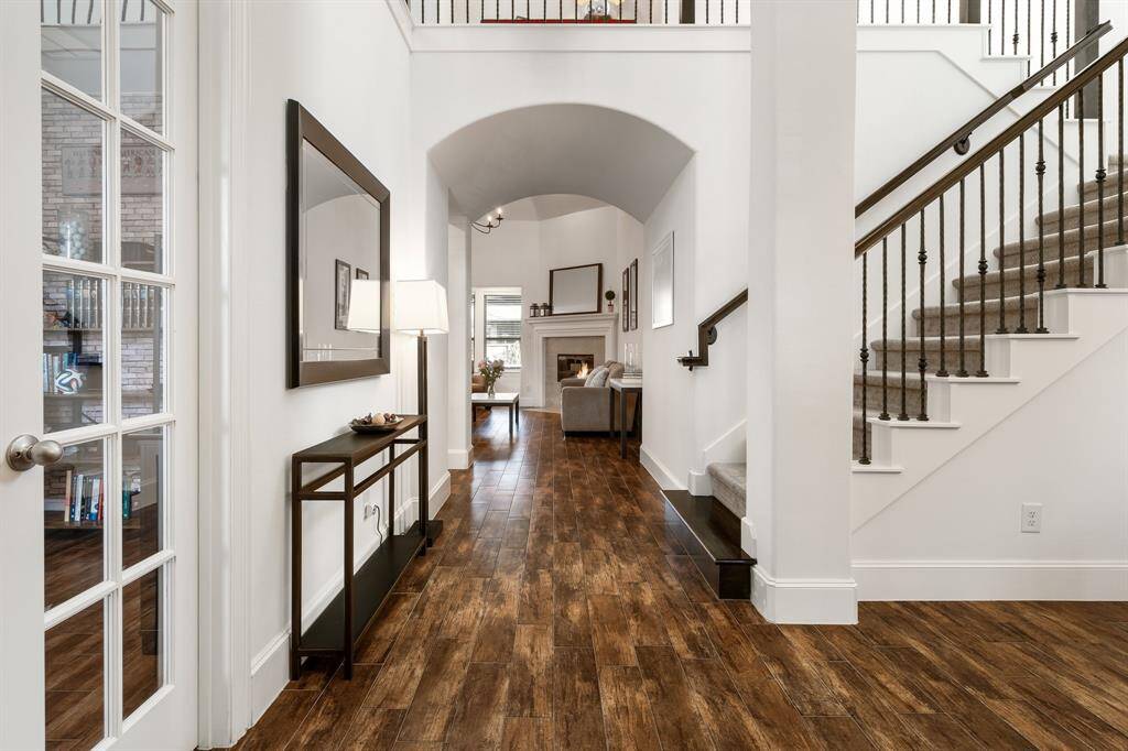 This inviting entryway features rich wood look like tile flooring, a spacious open hallway with high ceilings, and a view into a cozy living area with a fireplace. A staircase with elegant custom iron railing leads to the upper level, complementing the modern and warm design. The natural light from all the windows create an open airy vibe that pulls you in.