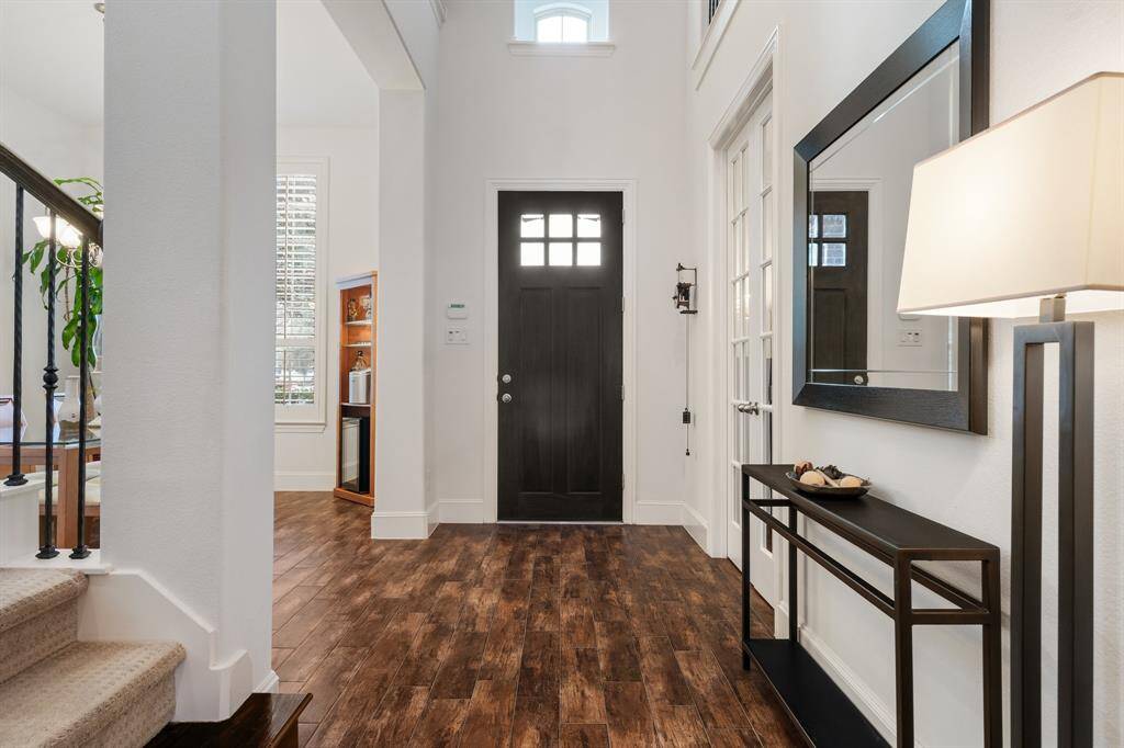 This entryway features a beautiful wooden front door with a window, leading into a bright, open formal dining room with hardwood look like tile floors. There's a stunning staircase on the left and an open foyer with soaring ceilings that leads to the study on the right of the entryway.