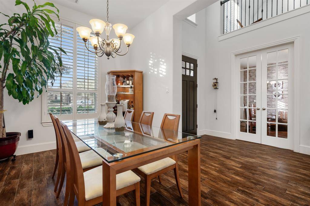 Bright formal dining area with a glass-top table, wooden chairs, and a large front window adorned with custom plantation wood shutters. Features a modern chandelier, look-like hardwood tile floors, and French doors leading to the study or 5th bedroom if you choose. High ceilings create an open and inviting atmosphere.