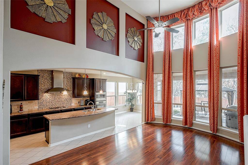 Spacious, open concept living area with two story high ceilings and large, bright windows features elegant red drapes, hardwood floors, and a stylish ceiling fan. The modern kitchen includes dark cabinetry, a tiled backsplash, and a granite countertop with pendant lighting.