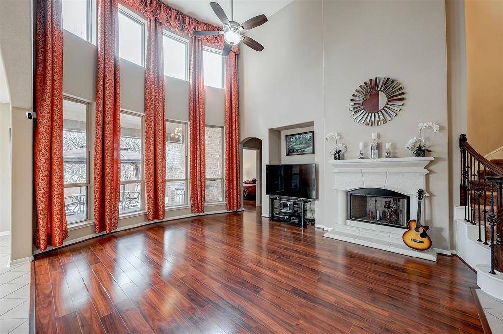 High ceilings and large windows with plenty of natural light. It has beautiful hardwood floors, a modern ceiling fan, and a stylish fireplace with decorative accents. The room also includes a TV area and an open view to a patio, creating an inviting and airy atmosphere.