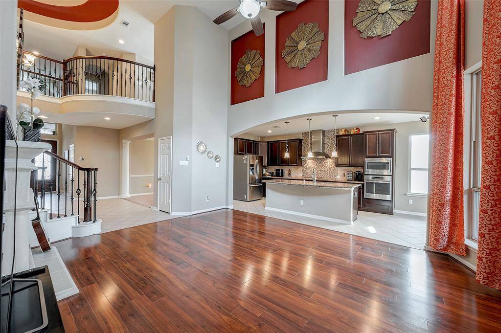 Another view to the open-concept living area features elegant wood flooring, high ceilings, and a modern kitchen with dark cabinetry and stainless steel appliances. The room is accented by large windows with red curtains, stylish wall decor, and a view of a sweeping staircase with wrought iron railings leading to an upper level.