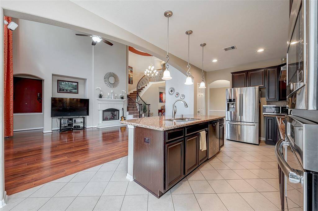 Elegant pendant lighting hangs over the kitchen island. The adjacent living space has rich wood flooring, a high ceiling with a fan, and a built-in fireplace, providing a warm and inviting atmosphere. A staircase leads to the upper level, adding architectural interest.