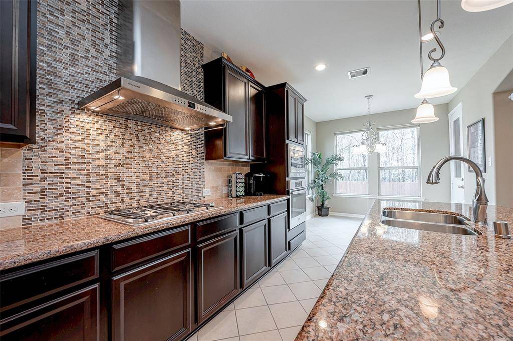 This modern kitchen features granite countertops, dark wood cabinets, and a stylish tile backsplash. It includes a gas cooktop with a stainless steel hood, a double sink, and pendant lighting. The space is bright and airy, with large windows offering plenty of natural light.