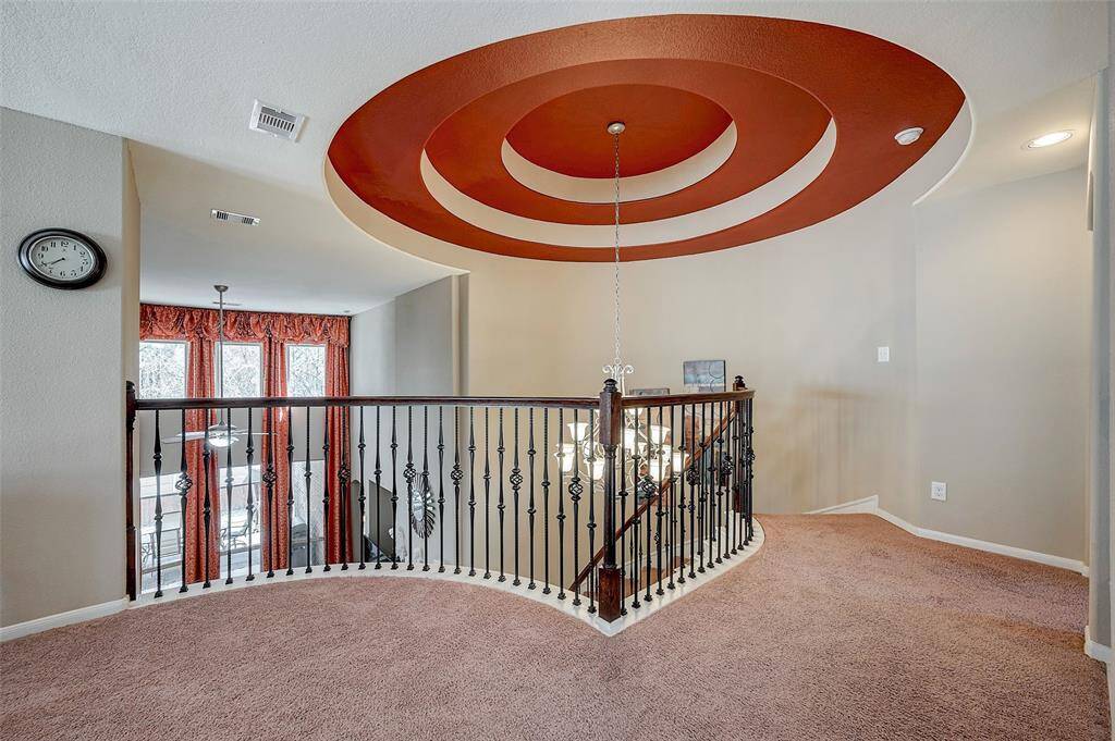 A stylish upper landing area with a striking circular ceiling design and a decorative chandelier. The space features a curved wrought iron railing, plush carpet, and ample natural light from large windows with red drapes, creating a warm and inviting atmosphere.