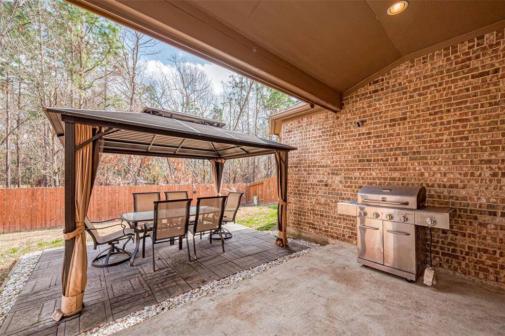 This backyard patio features a covered seating area with a table and chairs, perfect for outdoor dining. A brick wall provides a backdrop, and there's a built-in grill for barbecues. The space is bordered by a wooden fence and surrounded by trees, offering privacy and a natural setting.