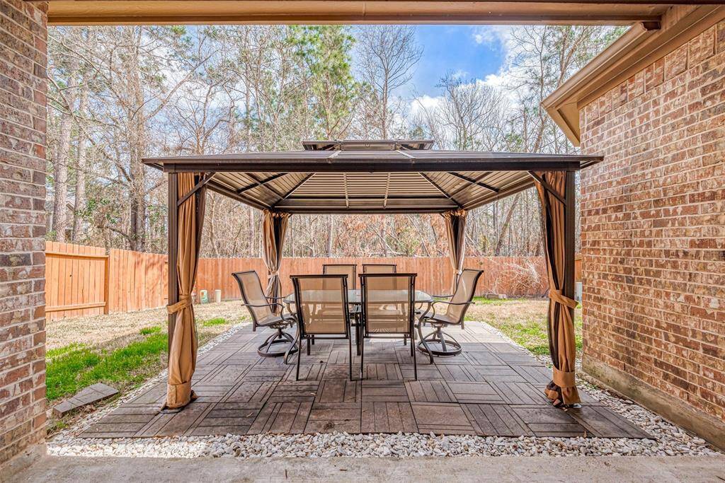 A cozy backyard patio featuring a covered gazebo with outdoor seating. It's surrounded by a wooden privacy fence and bordered by trees, providing a tranquil and private setting for relaxation or entertaining.