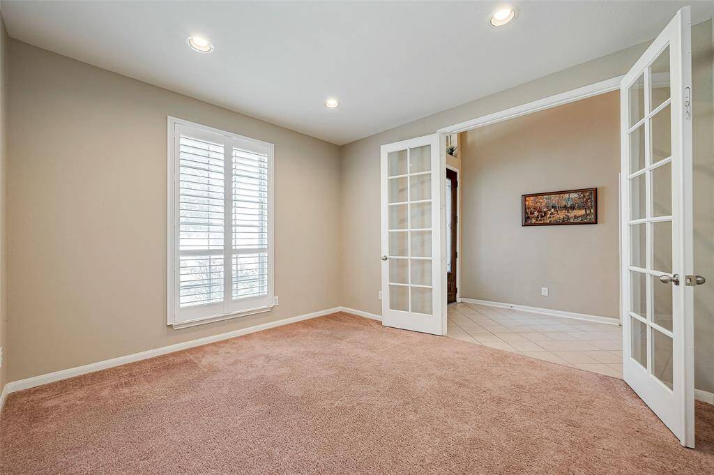 This office features a cozy carpeted floor, neutral walls, and ample natural light through a window with white shutters. It includes elegant French doors leading to an adjoining foyer creating an inviting and versatile space.
