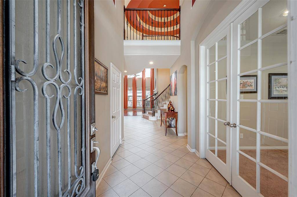 Entryway features a wrought iron wood door, tile flooring, and abundant natural light. The foyer gracefully leads to a staircase adorned with elegantly curved railings, while French doors opening into a home office or study.