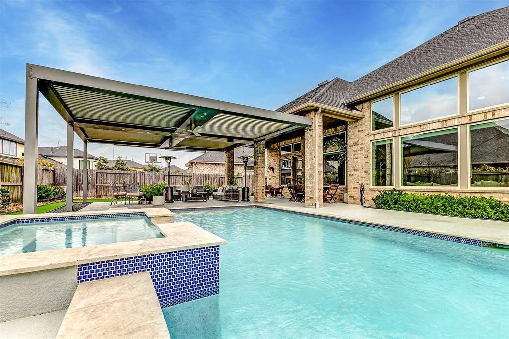 This photo showcases a beautiful backyard with a spacious pool and hot tub, complemented by a modern pergola-covered patio area. Large windows provide a view of the elegant brick house, creating a perfect setting for relaxation and entertainment.
