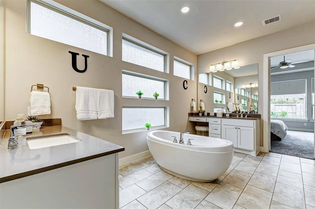 This modern bathroom features a sleek freestanding tub, dual vanities with ample counter space, and large frosted windows for natural light. The neutral color palette and contemporary fixtures create a serene, spa-like atmosphere, and the adjoining bedroom adds convenience.