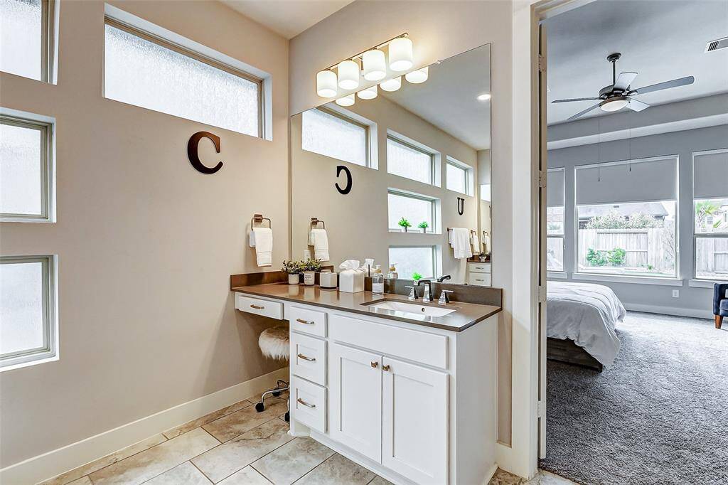 Sleek vanity with ample counter space, large mirror, and contemporary lighting. Frosted windows provide natural light while maintaining privacy. The adjoining bedroom is spacious, with plush carpeting, large windows, and a ceiling fan, offering a cozy and inviting atmosphere.