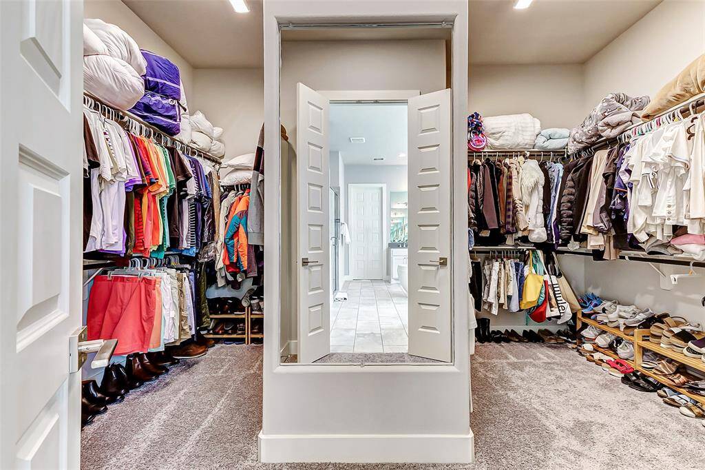 This is a spacious walk-in closet with ample shelving and hanging space, featuring a plush carpeted floor. It has neatly organized clothes, shoes, and accessories, and a doorway leading to a tiled bathroom area.
