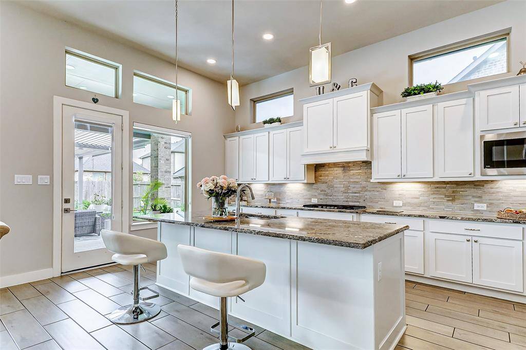 This modern kitchen features sleek white cabinetry, a spacious island with granite countertops, and contemporary pendant lighting. Large windows and a glass door provide ample natural light and access to a patio area. The space is accented with stainless steel appliances and a stylish backsplash.