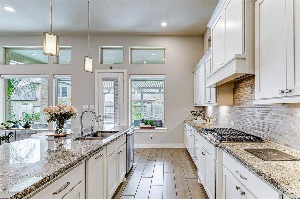 Bright, modern kitchen with granite countertops, white cabinetry, and stainless steel appliances. Features a central island with sink, pendant lighting, and large windows offering natural light and views of the backyard.