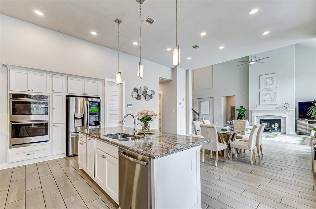 This spacious open-plan kitchen and living area features modern appliances, a large island with granite countertops, and elegant pendant lighting. It flows seamlessly into a bright dining and living space with a cozy fireplace, high ceilings, and ample natural light, creating a perfect atmosphere for entertaining and family gatherings.