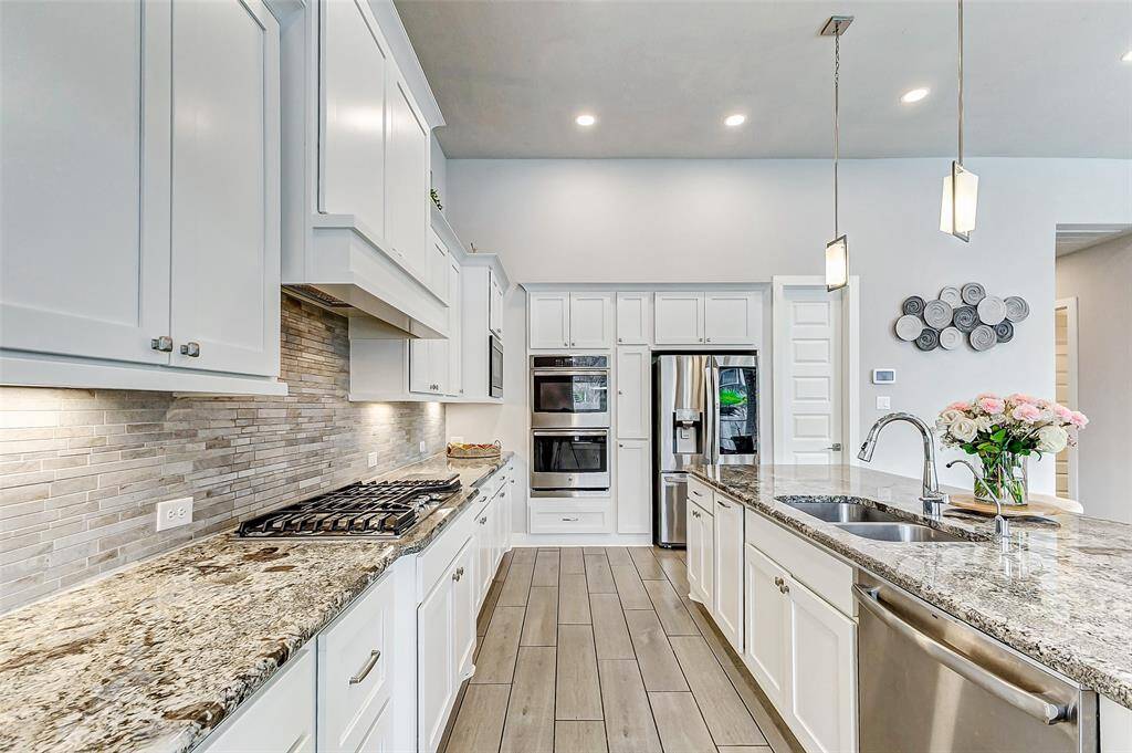 This modern kitchen features sleek white cabinetry, granite countertops, and stainless steel appliances. It includes a spacious island with a sink, pendant lighting, and a stylish tile backsplash. The open layout is complemented by wood-look tile flooring, creating an inviting and functional space.