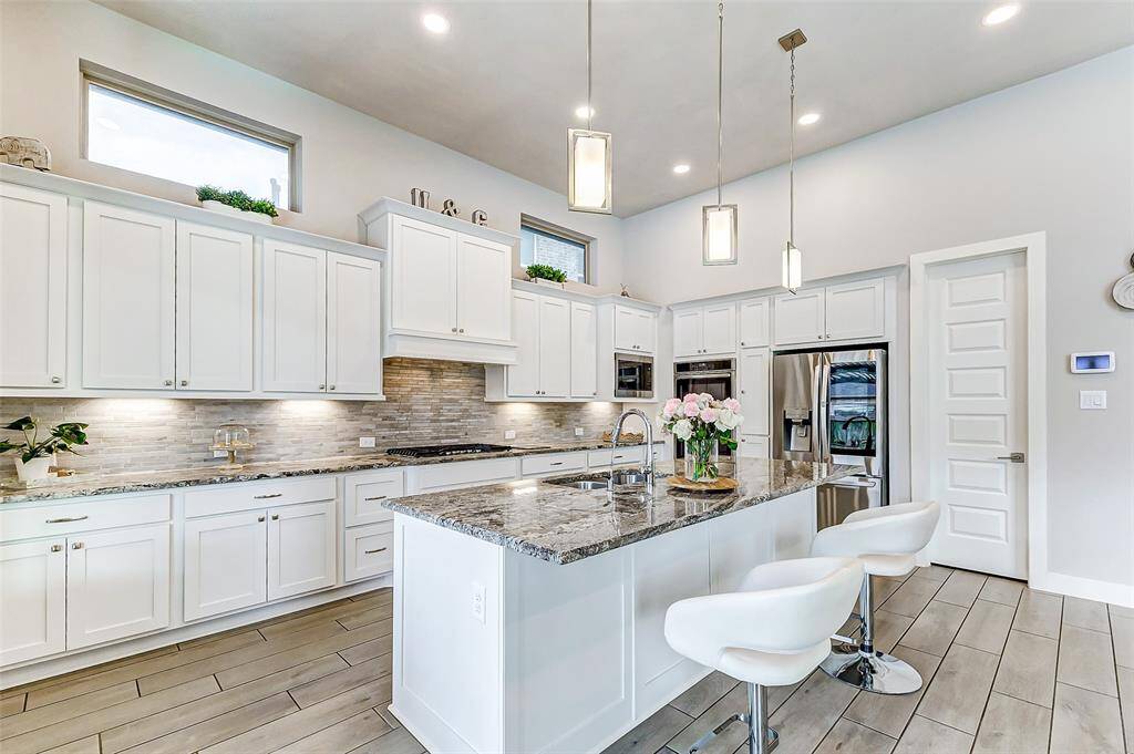 This modern kitchen features sleek white cabinetry, stainless steel appliances, and a large island with granite countertops. Pendant lighting enhances the bright, open space, complemented by stylish bar and a chic backsplash.