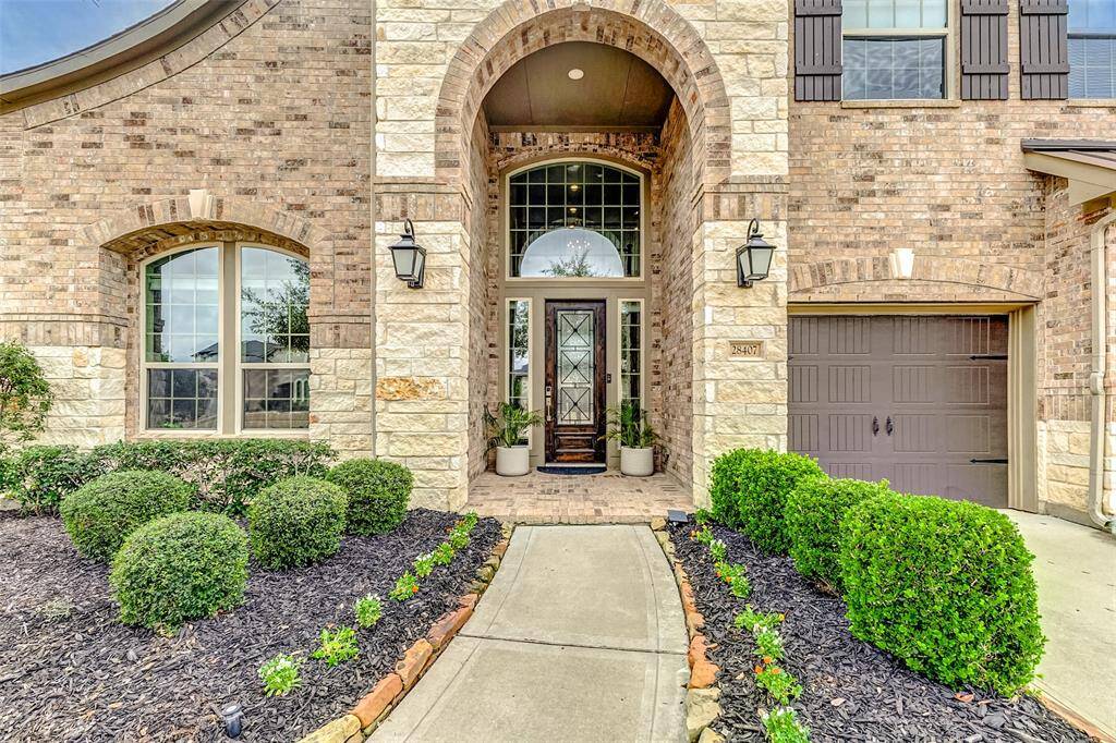 Elegant brick and stone home exterior with a welcoming arched entryway. The front door features glass panes, and the surrounding landscape is neatly manicured with shrubs and a tidy walkway leading to the entrance.