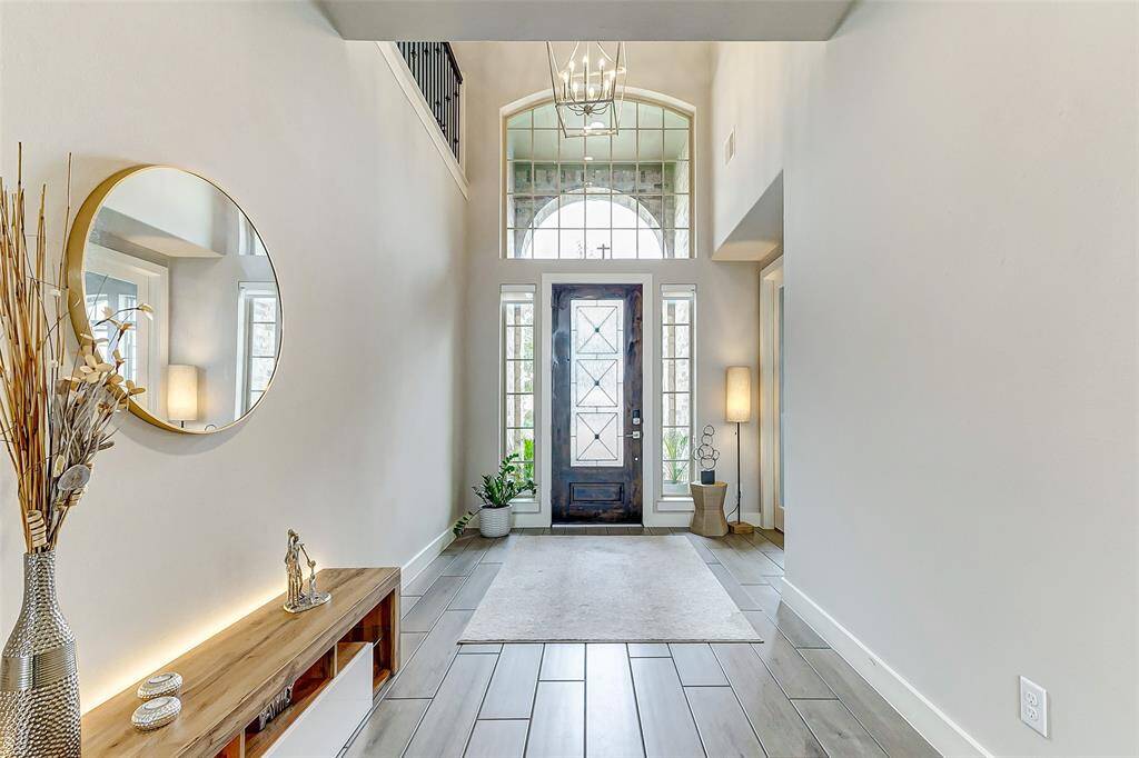 This entryway features a high ceiling and large windows around the front door, creating a bright and welcoming space. The modern decor includes a round mirror, decorative vase, and sleek bench, enhancing the contemporary feel.