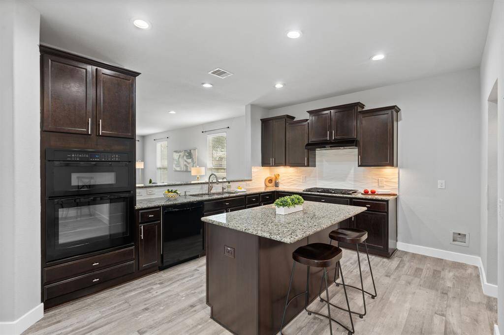 This modern kitchen boasts a large granite island, sleek cabinetry, and recessed lighting. The open layout seamlessly flows into adjacent rooms, with wood-style flooring enhancing the bright, airy feel. The high bar area offers a stylish division between the kitchen and gathering room, perfect for both functionality and desig