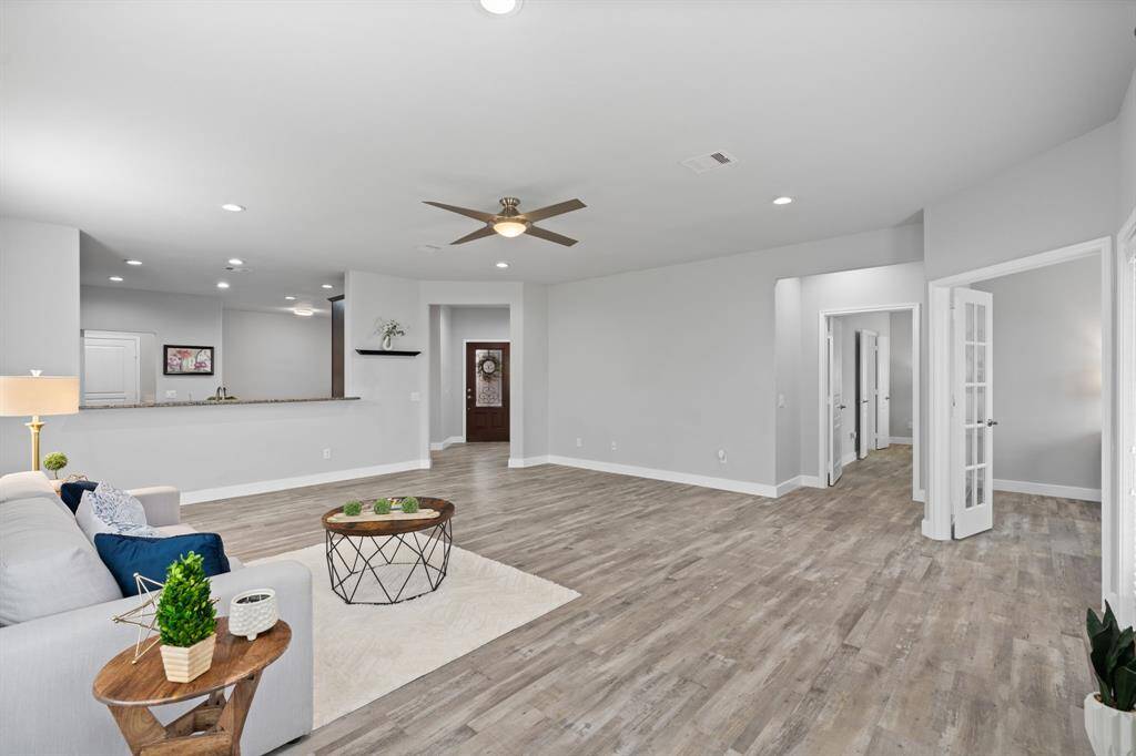 Spacious living area features an open floor plan with modern gray walls and wood-look flooring. It includes a ceiling fan, recessed lighting, and a cozy seating area. The room flows seamlessly into the kitchen with a breakfast bar, creating a perfect space for entertaining. French doors lead to additional rooms, enhancing the home's versatile layout.