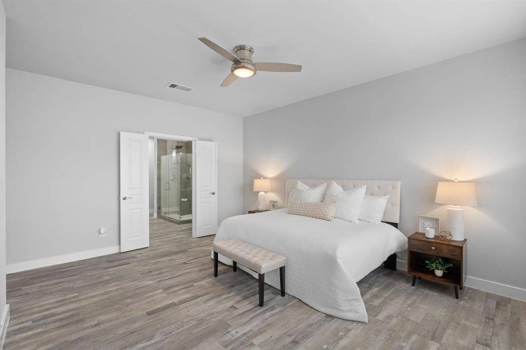 This bright and spacious bedroom features a king-sized bed, modern ceiling fan, and elegant wood flooring. It has an en-suite bathroom accessible through double doors, adding convenience and privacy. The room is tastefully decorated with neutral tones and soft lighting.
