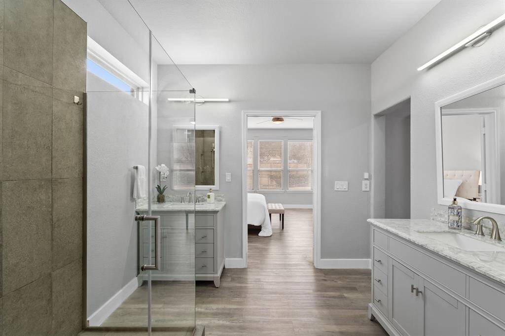 The owner’s bath has been beautifully remodeled into a modern retreat, featuring a glass-enclosed shower with striking dark gray penny round tile, contrasted by crisp white tile flooring. A double vanity with marble countertops, sleek gray cabinets, and framed mirrors adds elegance, while a statement light fixture enhances the sophisticated ambiance. The space is tied together with stylish wood-look flooring, creating a serene and contemporary design.