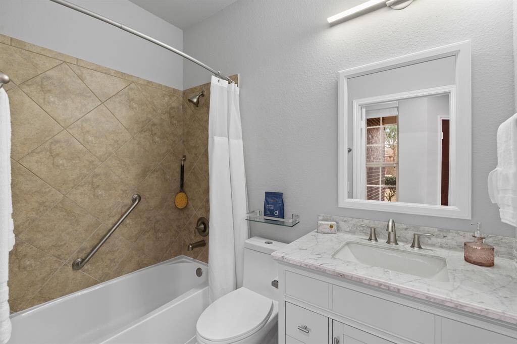 The guest bathroom showcases a fresh, modern update with a stylish tiled shower and tub combination, a sleek vanity with a marble countertop, and a framed mirror complemented by a unique light fixture. Even the toilet has been upgraded, ensuring a polished and contemporary feel throughout.