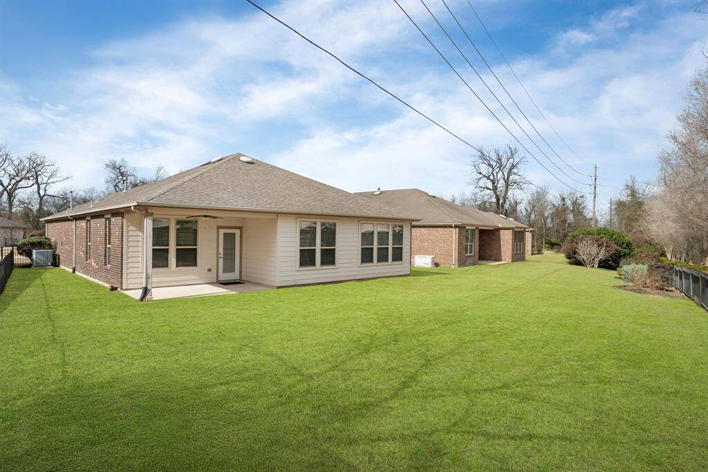 This photo shows a single-story brick house with a well-maintained backyard, featuring a spacious lawn and a small patio area. The property is fenced on one side and bordered by trees, offering privacy and a serene setting.