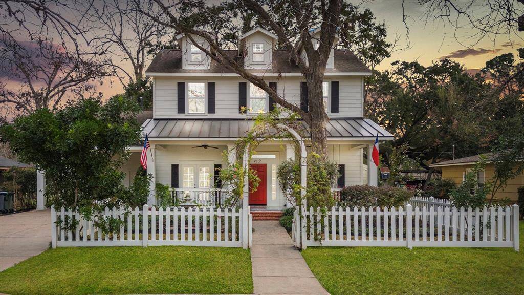 A beautiful white colonial house with a red door sits behind a picket fence, its entrance crowned by an arch of pink roses and greenery.