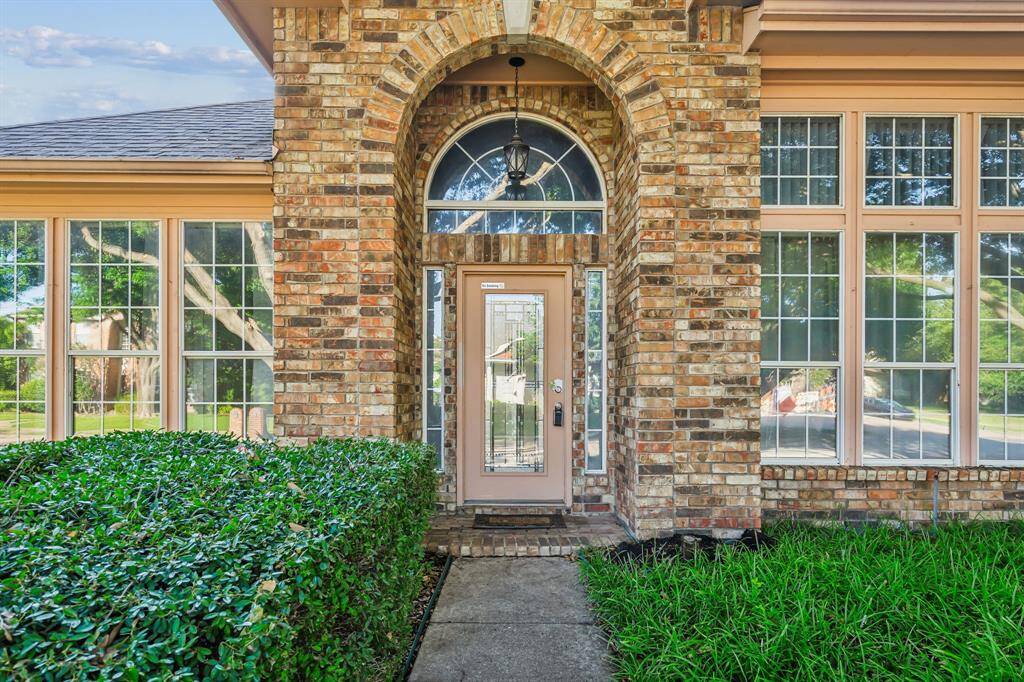 This photo showcases a welcoming brick home entrance with a decorative glass front door, framed by large windows and lush greenery. The arched design and well-maintained landscaping add charm and curb appeal.