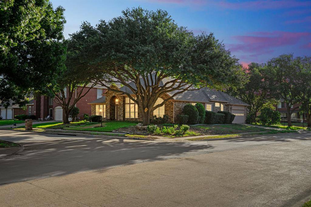 This photo showcases a charming, single-story brick home on a corner lot. The property features a well-maintained yard with mature trees and shrubs, and a two-car garage. The evening lighting highlights the welcoming exterior.