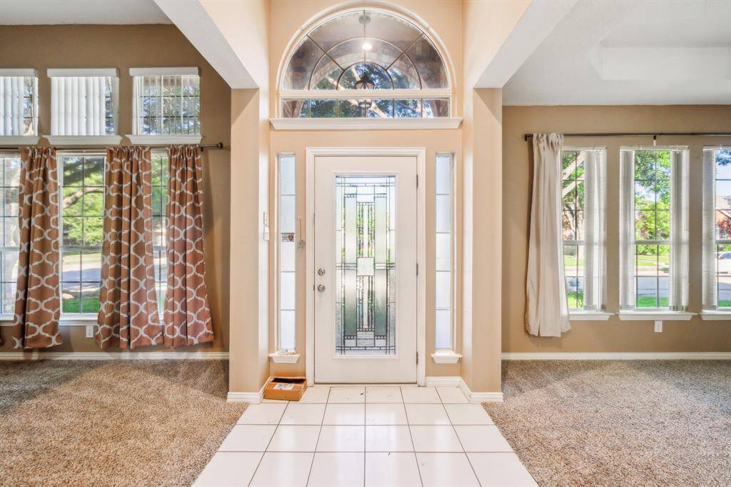This photo showcases a bright entryway with a decorative glass front door, flanked by tall windows that allow ample natural light. The space features tiled flooring leading into carpeted areas, and neutral walls provide a versatile backdrop.
