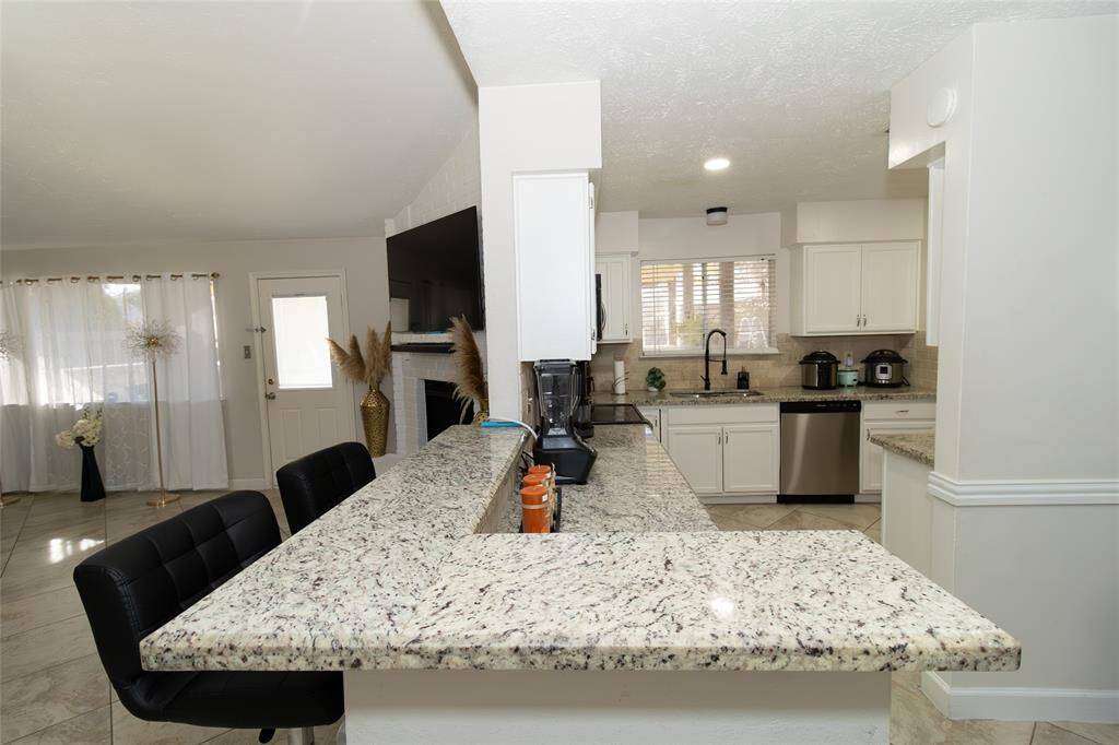 This photo showcases a modern kitchen with granite countertops, white cabinets, and stainless-steel appliances. There's a breakfast bar with seating, open to a bright living space with large windows.