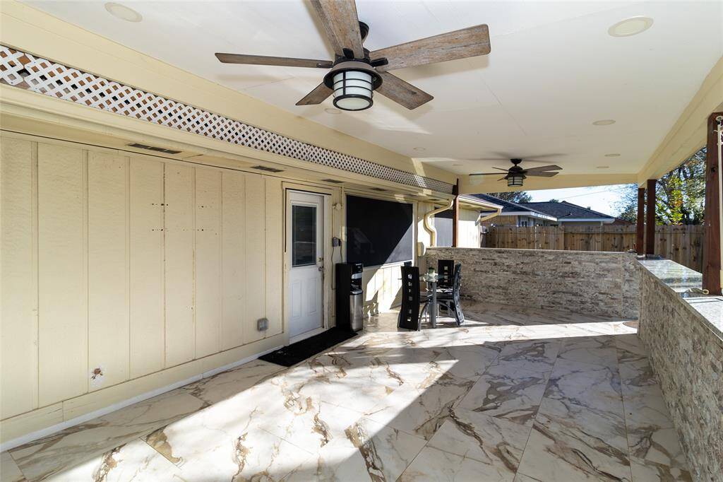 This photo showcases a spacious, covered outdoor patio with marble flooring and stylish ceiling fans. The area features a built-in countertop and seating, perfect for entertaining. A wooden fence provides privacy, while the ceiling and walls add a touch of elegance.