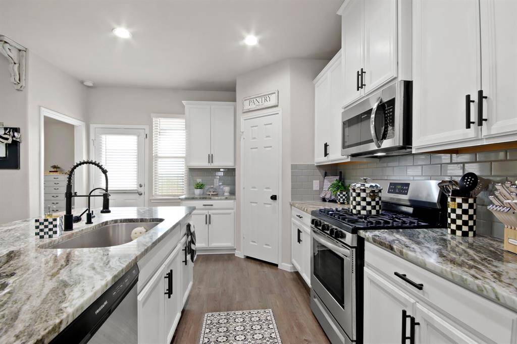 This kitchen features modern white cabinetry, stainless steel appliances, and sleek, granite countertops. The space includes a stainless sink with a stylish black faucet, a convenient pantry, and ample natural light from large windows. A chic backsplash and wood look vinyl flooring add to its contemporary appeal.