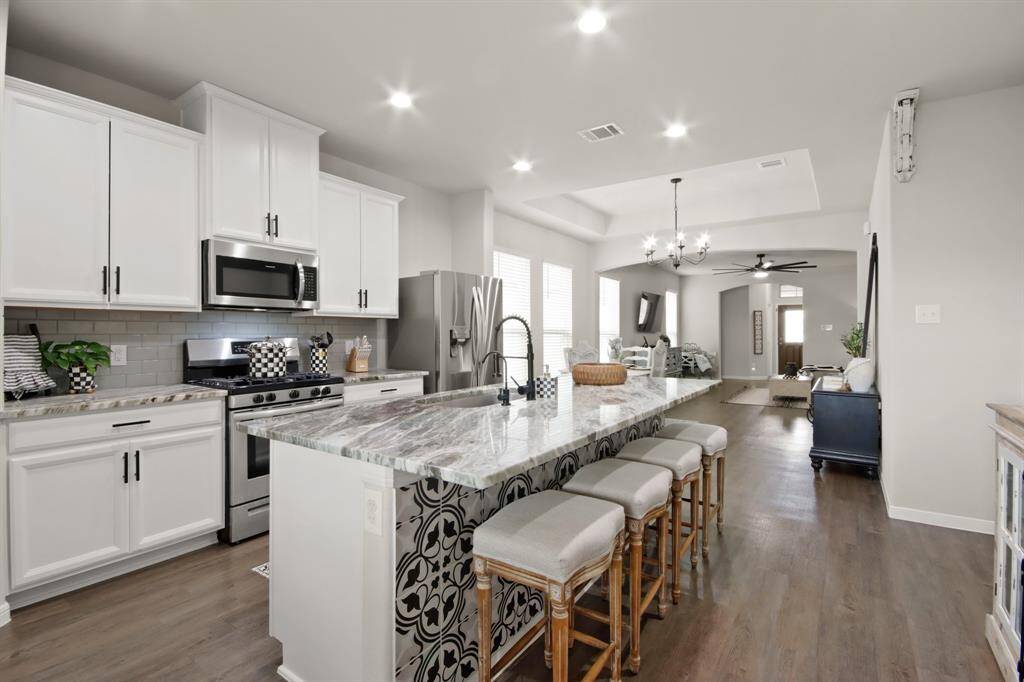 This photo showcases the modern, open-concept kitchen and living area with a large granite island with custom mosaic tile. The space is bright with recessed lighting, leading into a connected living room & dining room with a ceiling fan and chandelier.