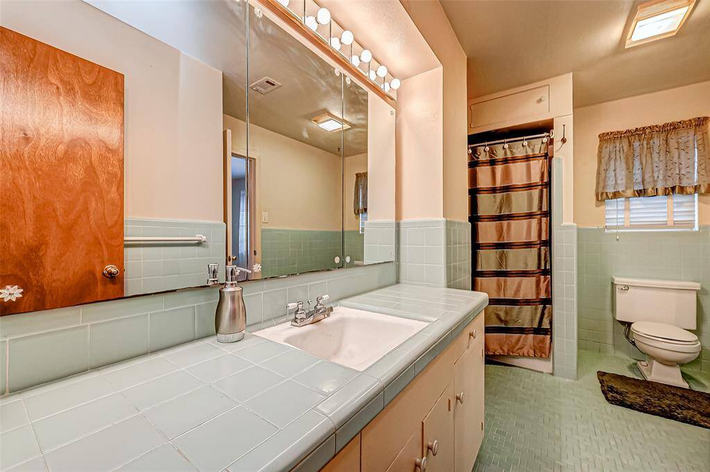 This bathroom features vintage green tile, a large vanity with ample counter space, and a full wall mirror with overhead lighting.