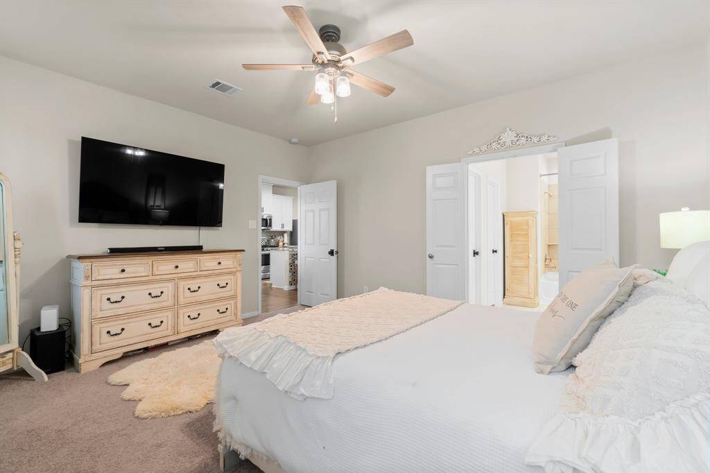 This bedroom is spacious and inviting, featuring an upgraded ceiling fan. The room has a neutral color palette and plush carpeting. It includes an en-suite bathroom with a visible tub, and there's a glimpse of a walk-in closet.