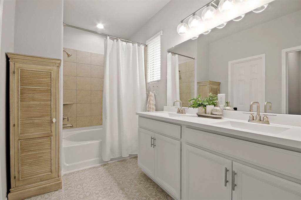 This bathroom features a double vanity with sleek modern fixtures, a large mirror, and ample counter space. It includes a tub-shower combination with a neutral color tile, complemented by decorative mosaic tiled flooring.
