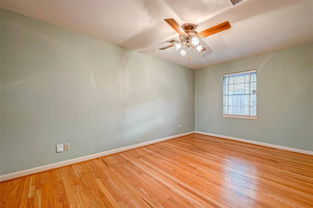 This is a bright, empty room with light hardwood floors and pale green walls. It features a ceiling fan with lights and a window allowing natural light.