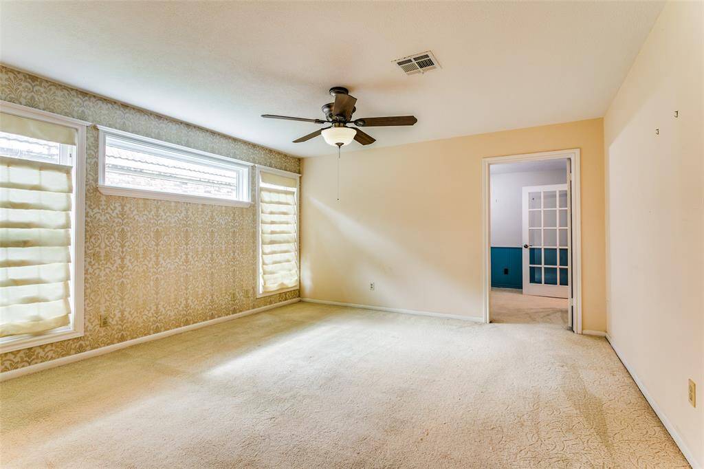 This is a bright, spacious primary bedroom featuring large windows with custom blinds, a ceiling fan, wall-to-wall carpeting, and a patterned wallpaper accent wall. The door leads to the adjacent study.