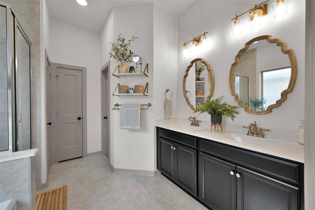 This is a modern and stylishbathroom featuring a double vanity, huge soaker tub and an enormous closet!with black cabinets and a whitecountertop, two unique gold framed mirrors above, and chicwall-mounted light fixtures.