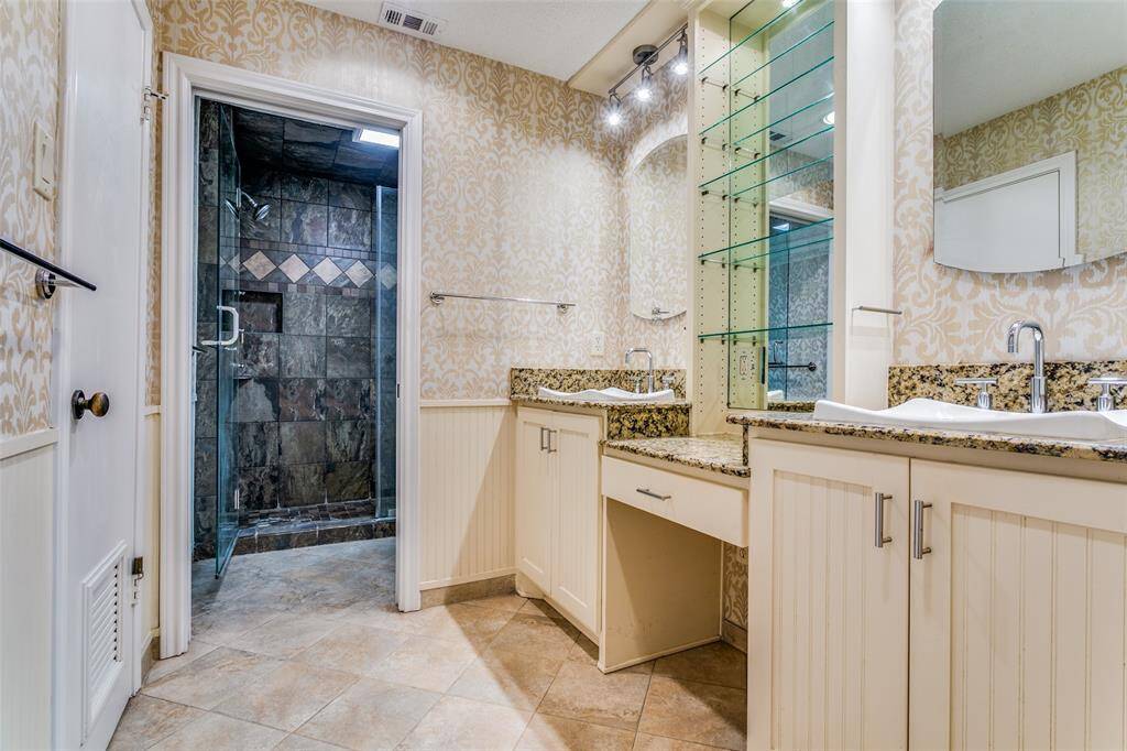 This spacious primary bathroom has a double vanity, granite countertops, updated sinks and faucets, and a large walk-in shower with custom tile work. It features ample lighting and a neutral color palette.