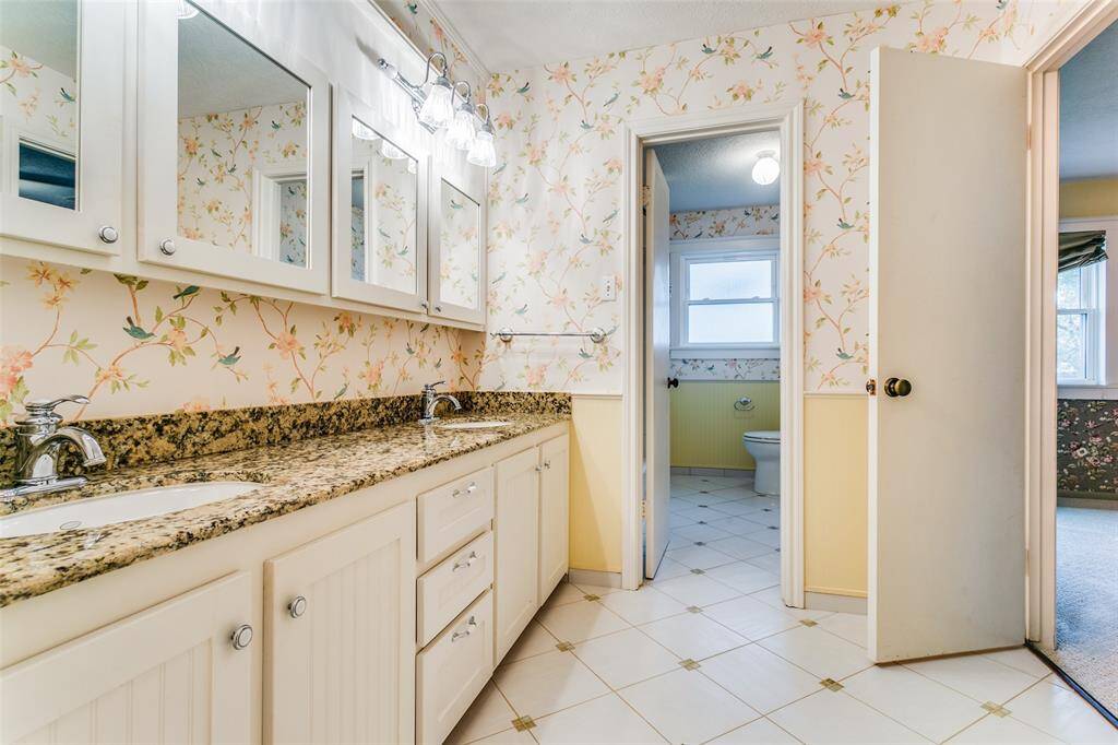 This brightly lit bathroom features a large vanity with granite countertops, updated sinks and faucets, tiled flooring with accents, floral wallpaper and a separate toilet area with window.