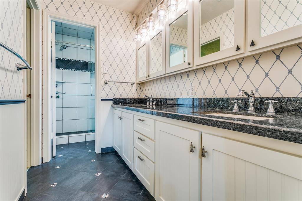 This is a clean, bright bathroom with granite countertops, updated sinks and faucets, tiled flooring with accents, updated lighting and separate shower with glass door and toilet.